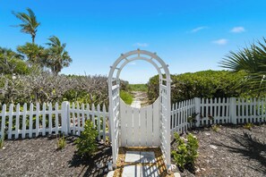 A white gate with a circular archway leading to a pathway surrounded by white picket fences and greenery. - 5BR Beachfront Escape | Private Beach Access (Cape Canaveral)