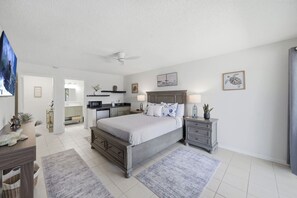 Une chambre à coucher comprenant un lit, des tables de chevet, une commode, un ventilateur de plafond et donnant sur la cuisine.