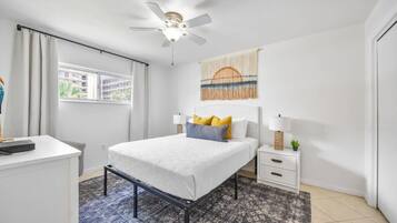 Une chambre à coucher avec un lit, des tables de chevet, un ventilateur de plafond, une fenêtre et une vue sur les bâtiments environnants.