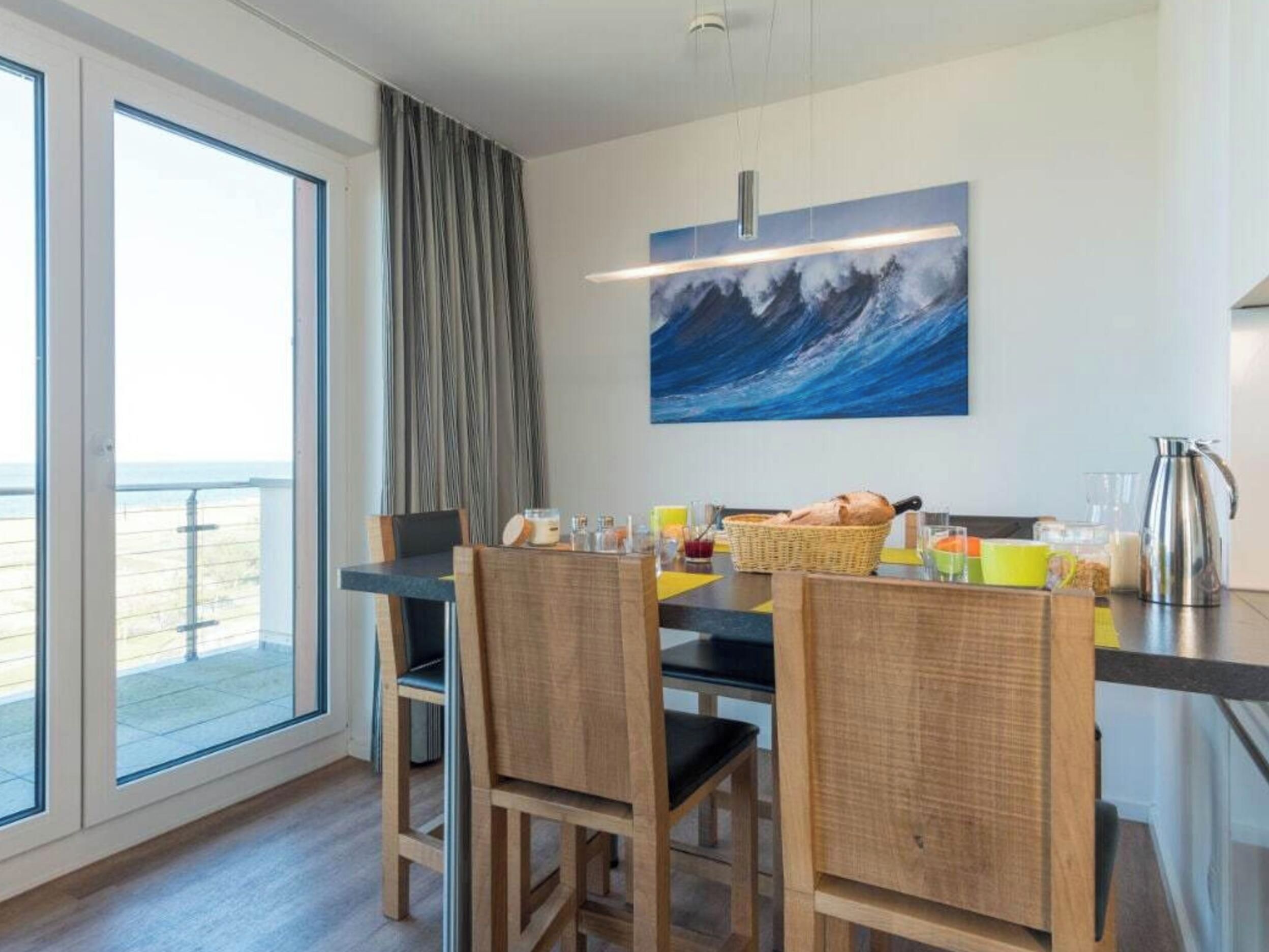 Uma sala de jantar com vista para o mar, um quadro na parede e itens de café da manhã sobre a mesa.