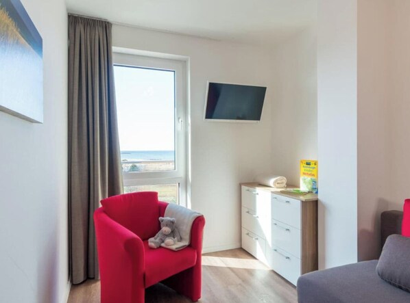 A room with a red chair, a grey sofa, a television on the wall, a window with a view, and a small cabinet with a towel.