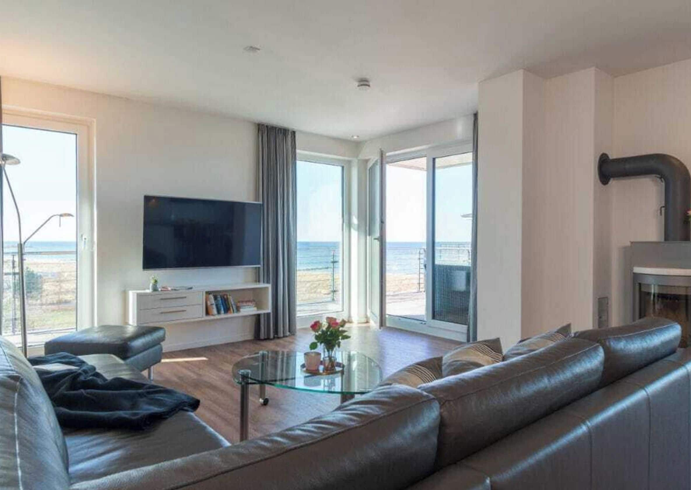 A modern living room with a large flat-screen TV, leather sofas, a glass coffee table, and a view of the sea through large windows.