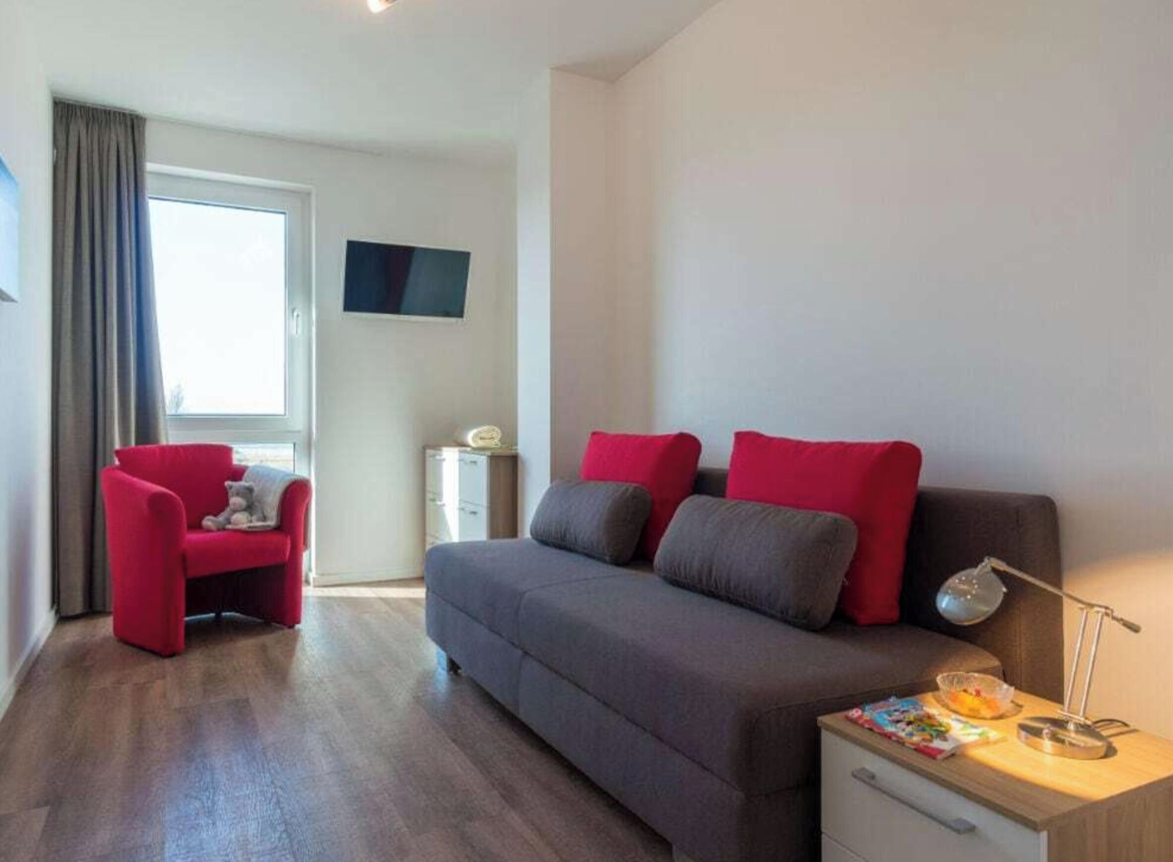 A modern bedroom with a grey sofa, red cushions, a red armchair, a bedside table with a lamp, and a TV mounted on the wall.