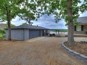 A residential garage with a gravel driveway, a covered outdoor seating area with chairs, and a view of a body of water. - Charming lake front 2-bedroom cottage with AC on peaceful Lake Eufaula, OK. (Canadian)