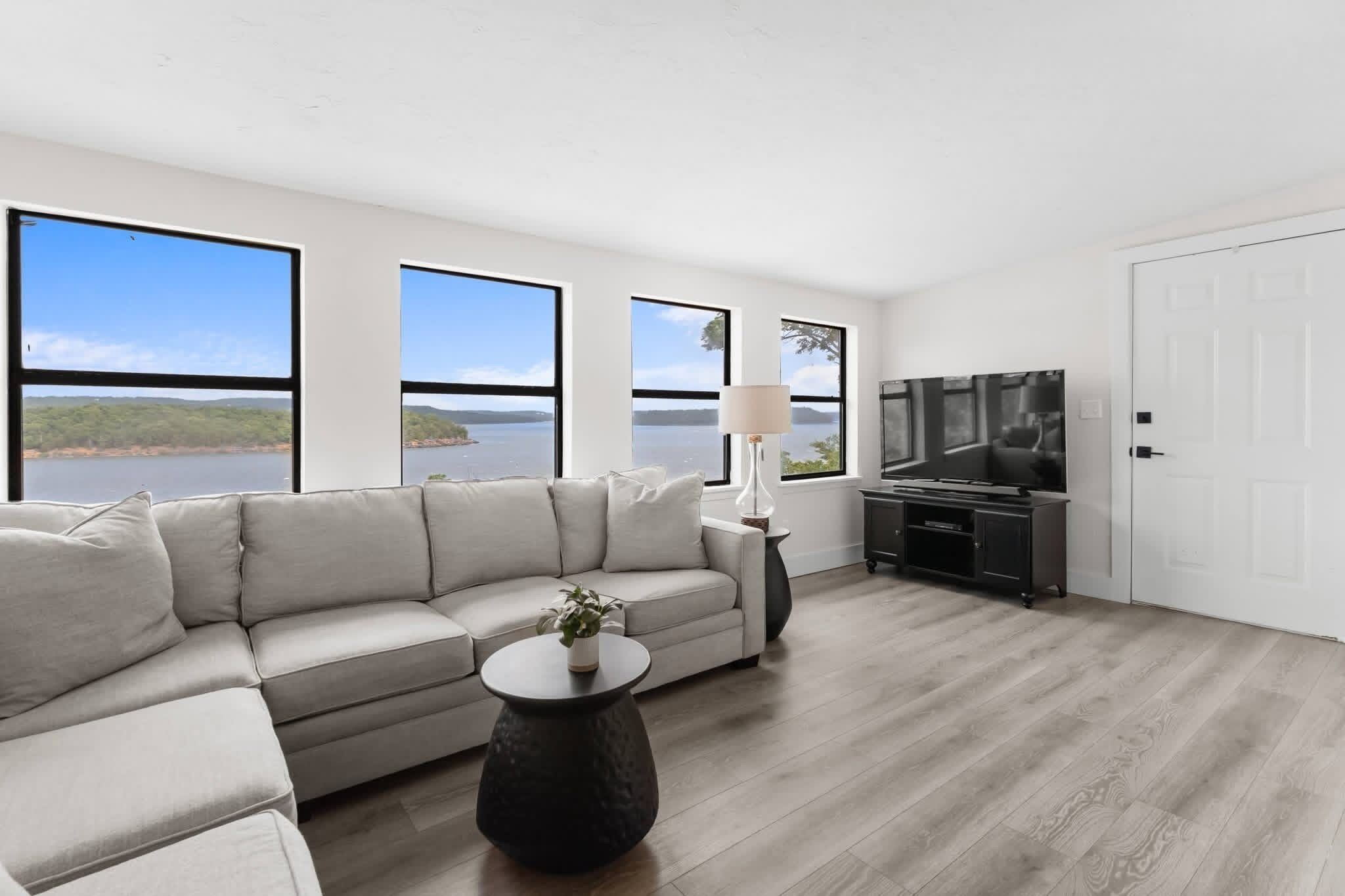 Una sala de estar moderna con un sofá, una mesa de centro, un televisor y ventanas amplias con vistas a un lago.