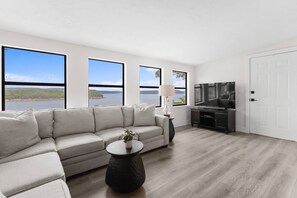 A modern living room with a sofa, a coffee table, a TV, and large windows offering a view of a lake. - Charming lake front 2-bedroom cottage with AC on peaceful Lake Eufaula, OK. (Canadian)