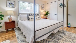 A bedroom with a four-poster bed, a bedside table with a lamp, a potted plant, and artwork on the walls. - A-Wave From It All #1 - Pet-Friendly Pool Home (New Smyrna Beach)