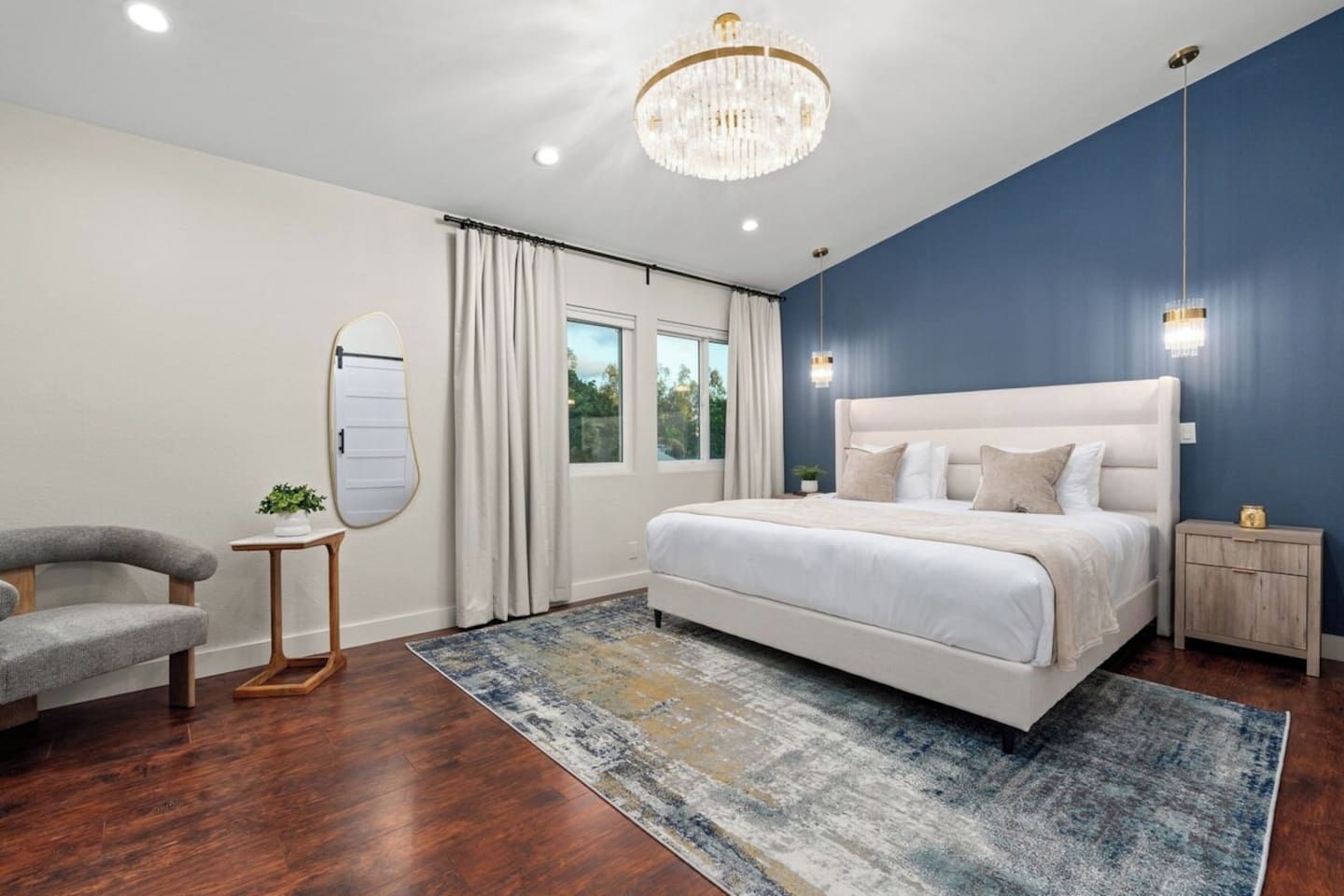 A bedroom with a large bed, a chandelier, a nightstand, a chair, and a window with curtains.