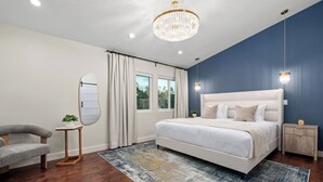 Ein Schlafzimmer mit einem großen Bett, einer Chandelier, einem Nachttisch, einem Sessel und einem Fenster mit Vorhängen.