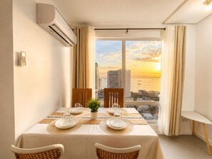 Zona pranzo con un tavolo apparecchiato per due, una pianta in vaso e vista sulla città al tramonto.