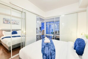 A modern hotel room with a large bed, a desk, and a view of the city through a glass wall. - Comfort 1BR Hotel Suite at The Velaire Toronto | Sleeps 4 (Toronto)