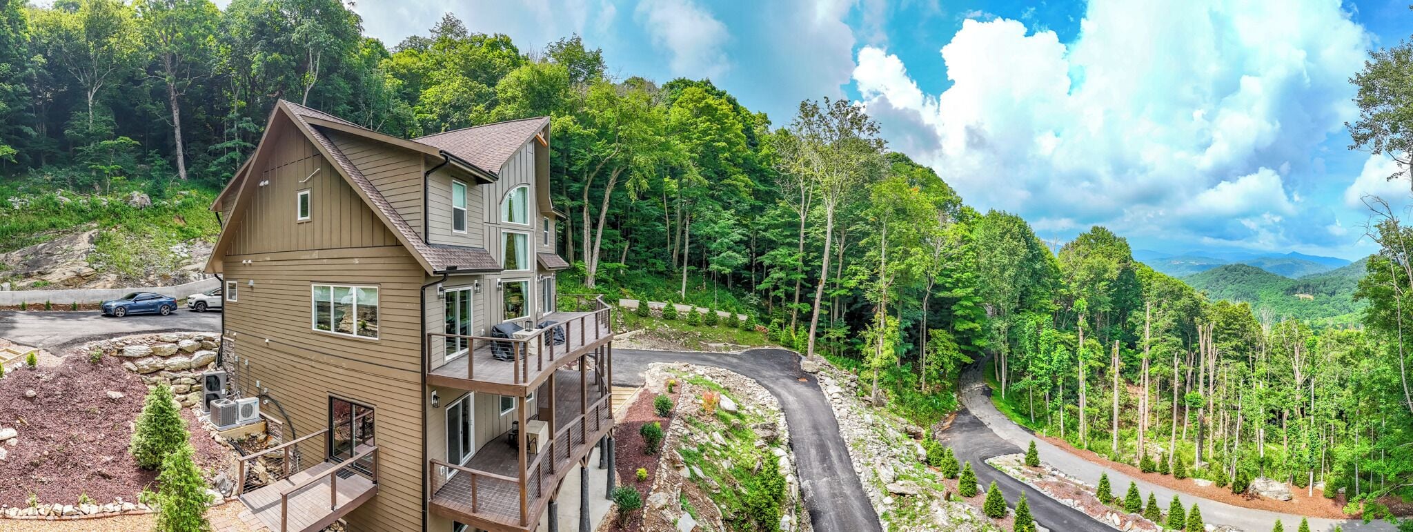 A multi-story house with a balcony, surrounded by a forested landscape and a winding road.