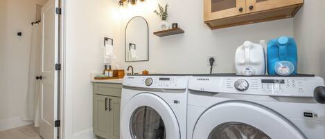 A laundry room with two front-loading washing machines, a sink, a mirror, and a shelf with toiletries.