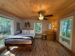 A bedroom with a wooden ceiling, a bed, a nightstand, a lamp, and large windows. - Loon Cabin – Gichigami Island Properties (La Pointe)