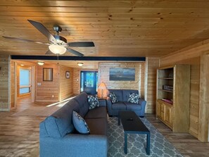 A log cabin interior with a sectional sofa, a coffee table, a ceiling fan, and a fireplace. - Loon Cabin – Gichigami Island Properties (La Pointe)