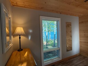 A room with a wooden floor, a window with a view of trees and a blue building, and a lamp on a wooden table. - Loon Cabin – Gichigami Island Properties (La Pointe)