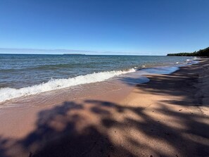 A beach with a clear sky, waves gently lapping at the shore, and a line of trees casting shadows on the sand. - Loon Cabin – Gichigami Island Properties (La Pointe)