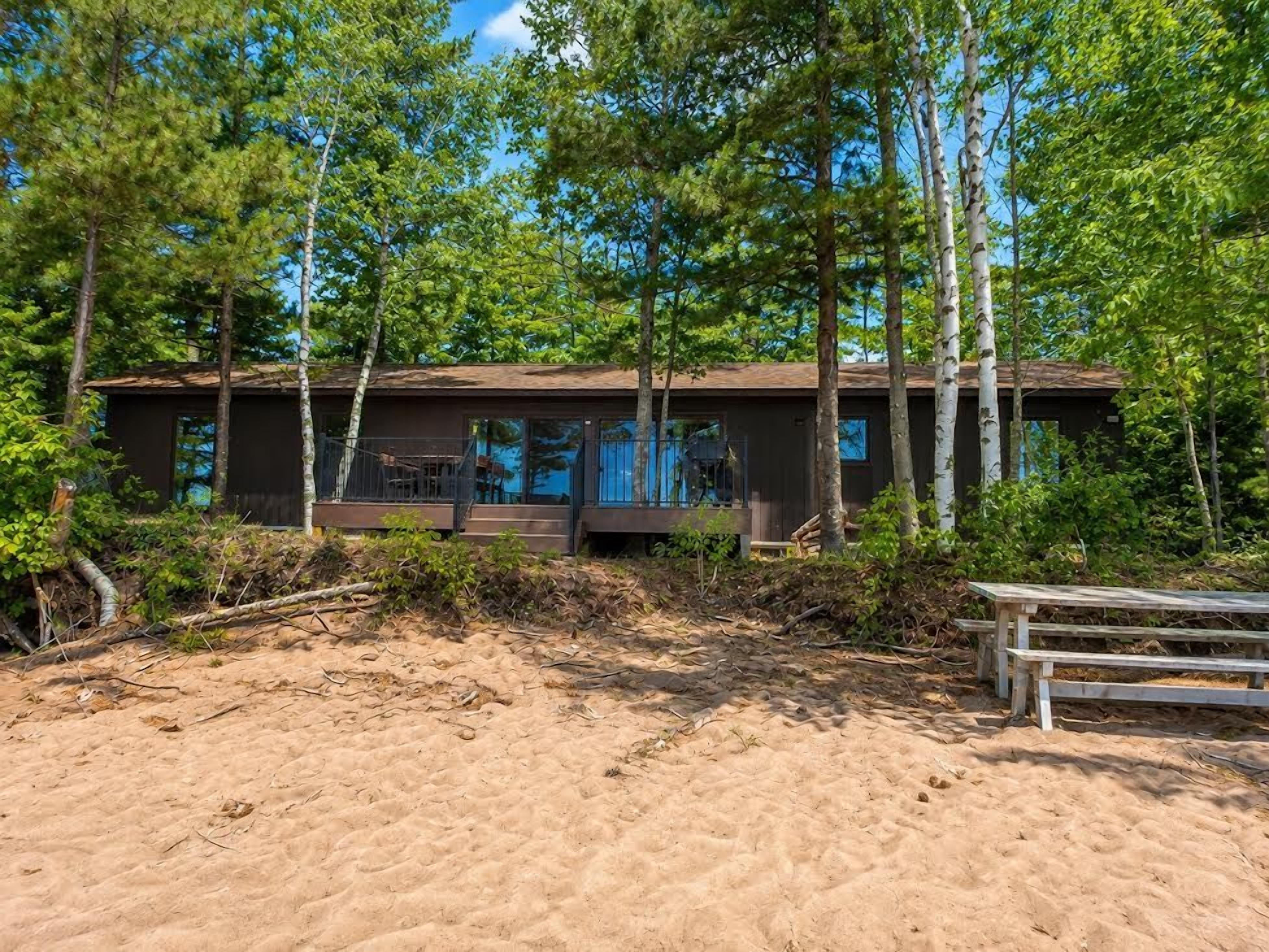Eine moderne Holzhütte mit einer Terrasse, umgeben von Bäumen und einem Sandstrand.