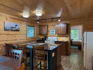 A wooden cabin interior with a kitchen area, dining table, chairs, and a refrigerator. - Loon Cabin – Gichigami Island Properties (La Pointe)