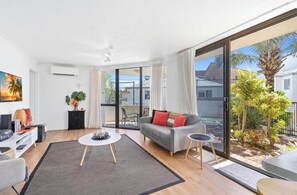 A modern living room with a sofa, coffee table, and a TV. There is a sliding glass door leading to a balcony with plants. - Coastal Retreat | Beach, Pool & Tennis (Palm Beach)