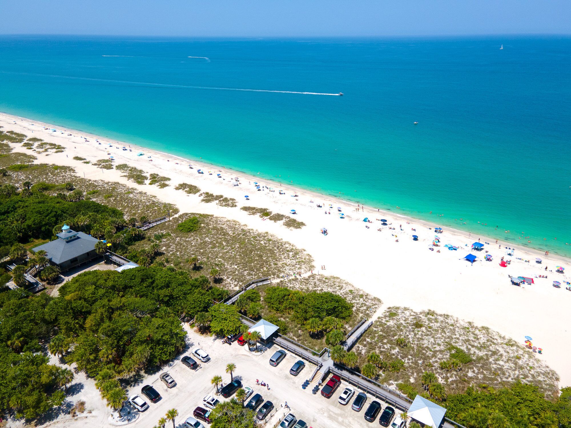 An aerial view of a beach with a parking area, beach umbrellas, and a clear view of the ocean.