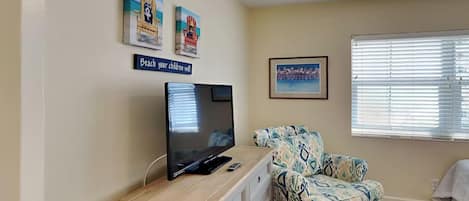 Una habitación con una televisión de pantalla plana sobre un mueble de madera, un sofá estampado, un cuadro enmarcado en la pared y una ventana con persianas.
