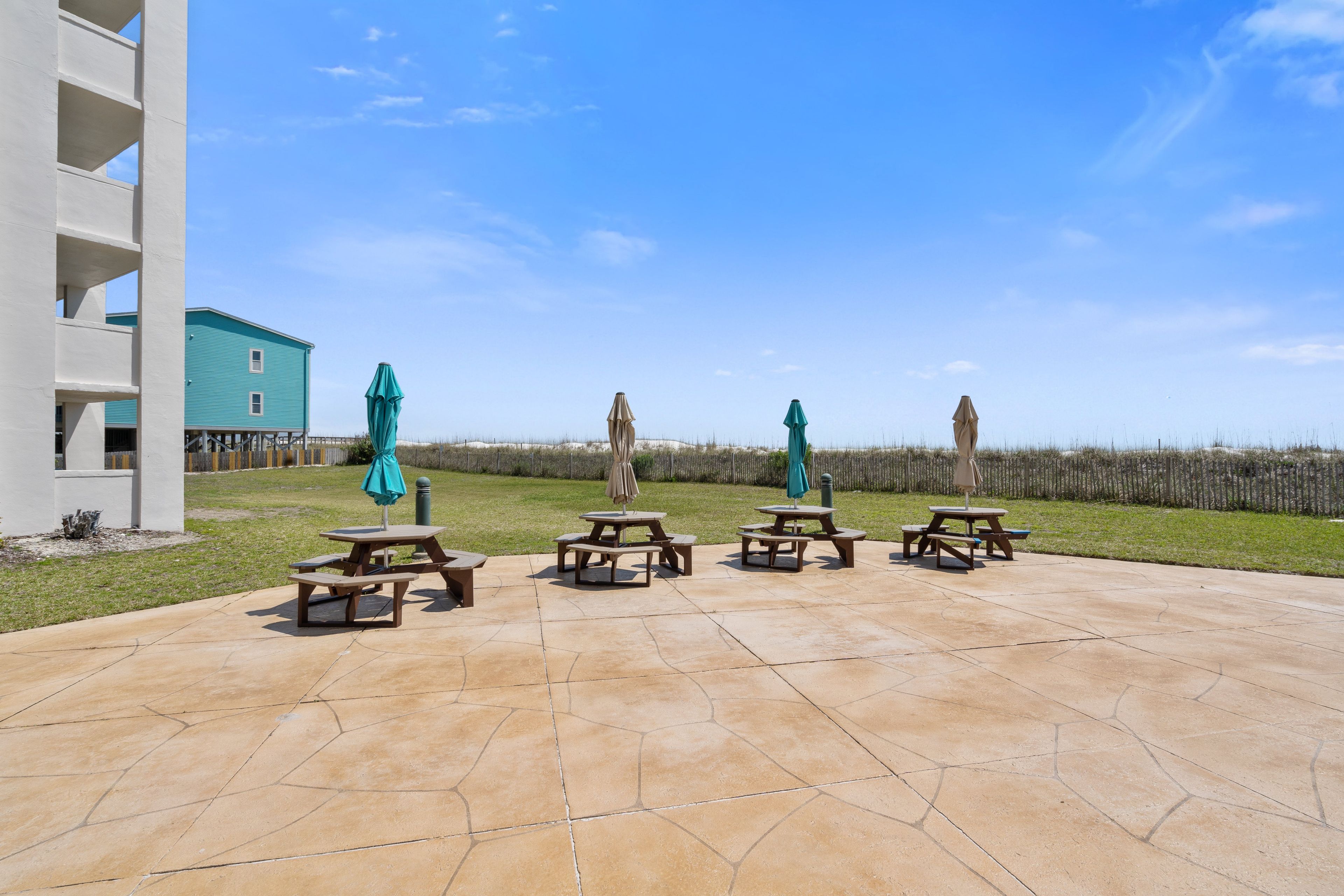 A patio with picnic tables and umbrellas, a building with a blue exterior, and a grassy area with a fence in the background.