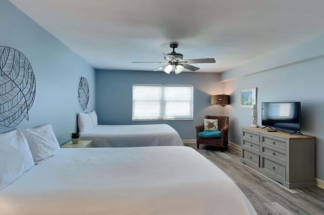 A hotel room with two beds, a ceiling fan, a TV, a chair, and a window with blinds.