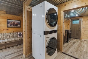 A compact laundry room with a stacked washer and dryer, wooden walls, a sofa with patterned cushions, and a framed picture on the wall. - Welcome to the Sheriff's Office (Stanton)