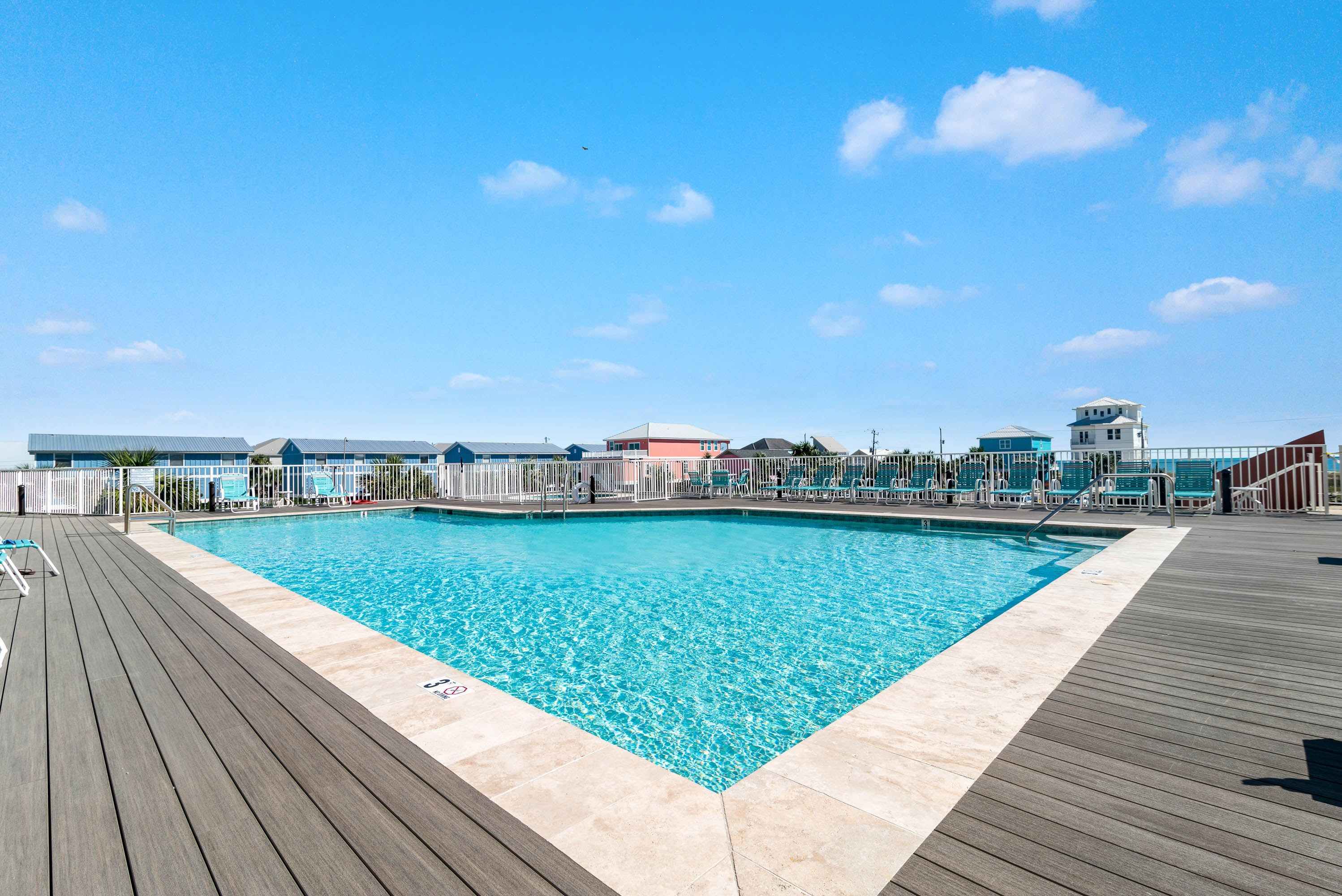 A swimming pool with a wooden deck and lounge chairs, surrounded by buildings with a clear blue sky above.