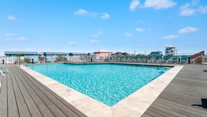 Ein Schwimmbecken mit Holzterrasse und Liegestühlen, umgeben von Gebäuden, mit blauem Himmel darüber.