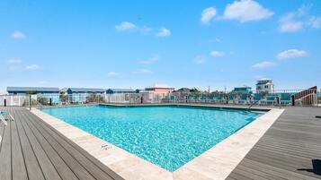 A swimming pool with a wooden deck and lounge chairs, surrounded by buildings with a clear blue sky above.