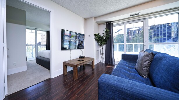 Ein Wohnzimmer mit einem blauen Sofa, einem Holztisch, einem Flachbildfernseher und einem großen Fenster mit Blick auf Gebäude.