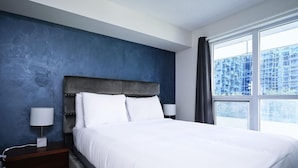 Ein Schlafzimmer mit einem großen Bett, einem Nachttisch, einer Lampe und einem Fenster mit Blick auf Gebäude.