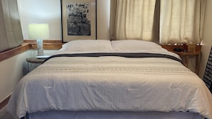 Een hotelkamer met een groot bed, een nachtkastje met een lamp, een lijst met een afbeelding aan de muur en een raam met gordijnen.