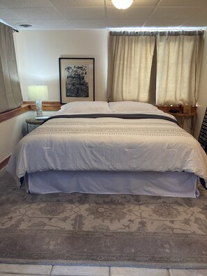A hotel room with a large bed, a bedside table with a lamp, a framed picture on the wall, and a window with curtains. - Charming 3-bedroom house on Fife Lake perfect for enjoying any season (Fife Lake)