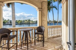 A balcony with wicker chairs and a small round table, overlooking a body of water with palm trees and buildings in the background. - The Palazzo | Waterfront | Walk to Beach | Luxury Villa (Fort Lauderdale)