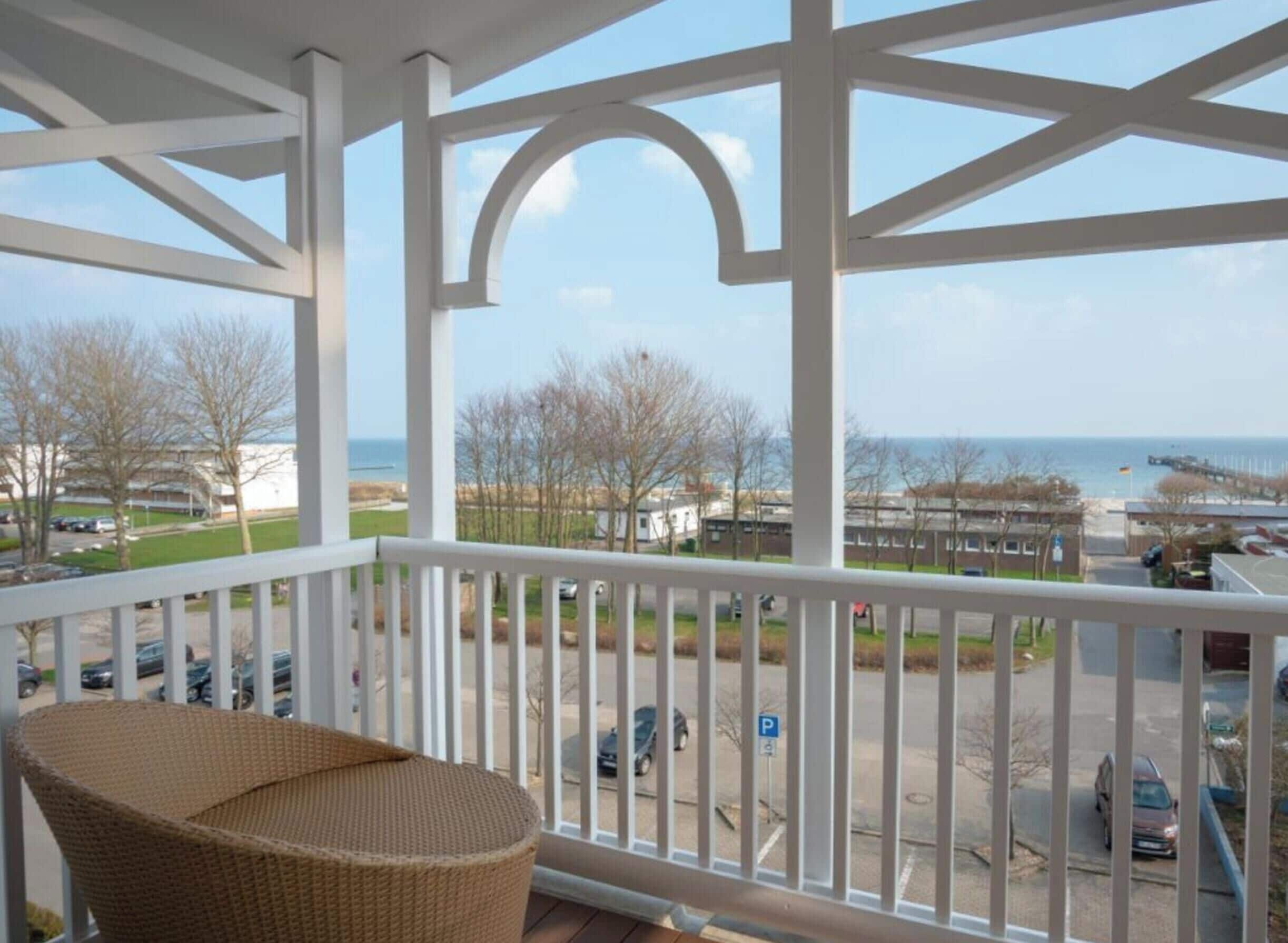 A balcony with a wicker chair, white railings, and a view of the sea and buildings.