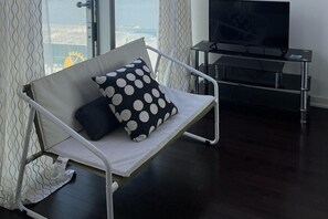 A modern living room with a white sofa, a black and white polka dot pillow, a flat-screen TV on a stand, and a view through large windows. - Basic Queen Studio Suite at The Velaire Toronto (Toronto)
