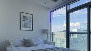 Ein modernes Hotelzimmer mit einem großen Bett, einem gerahmten Bild an der Wand, einem Nachttisch mit Lampe und Blick auf die Stadt durch ein großes Fenster.