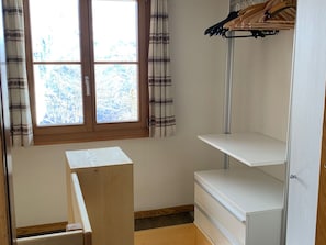 Ein Zimmer mit Fenster, einem Holzschrank, einer weißen Regaleinheit und einem Kleiderständer.