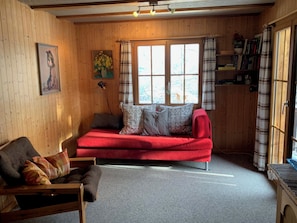 A cozy room with a red sofa, a wooden chair, a bookshelf, and a large window with curtains.