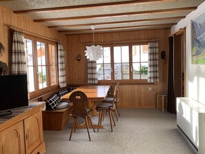 A wooden interior with a dining table, chairs, a television, and a window with curtains.