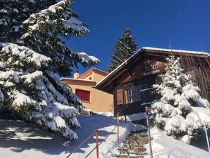 A wooden chalet with a red door, surrounded by snow-covered trees, under a clear blue sky.