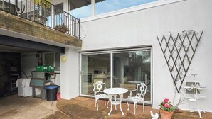 A residential property with a patio area featuring a table and chairs, a sliding glass door leading to a kitchen, and a balcony with a railing. - Apartment Sleeps 2 with Parking, Pets Welcome (Penrith)