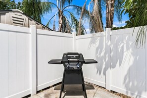 Una parrilla exterior negra con pantalla digital, ubicada en un patio trasero con cercas de madera blanca y palmeras al fondo.