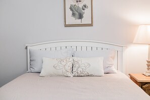Una cama con cabecera blanca, dos almohadas e ilustración botánica enmarcada en la pared.