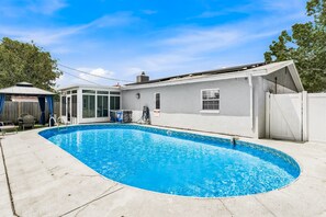 A residential swimming pool with a curved edge, a covered patio area, and a white house with a garage. - 🌸Pinellas Paradise - 4BR Pinellas Park home with pool| Sleeps 10 🌸 (Pinellas Park)