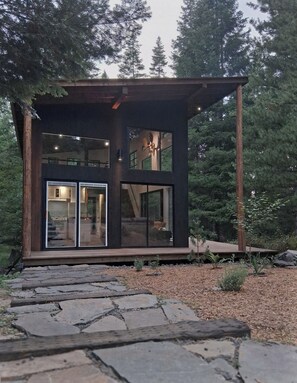 A modern cabin with large glass doors, a stone pathway leading to it, and a forested backdrop.