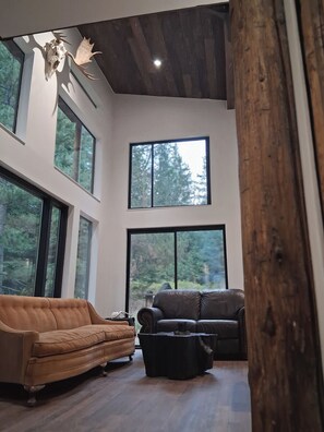 A modern living room with large windows, a brown sofa, a black ottoman, and a wooden ceiling.